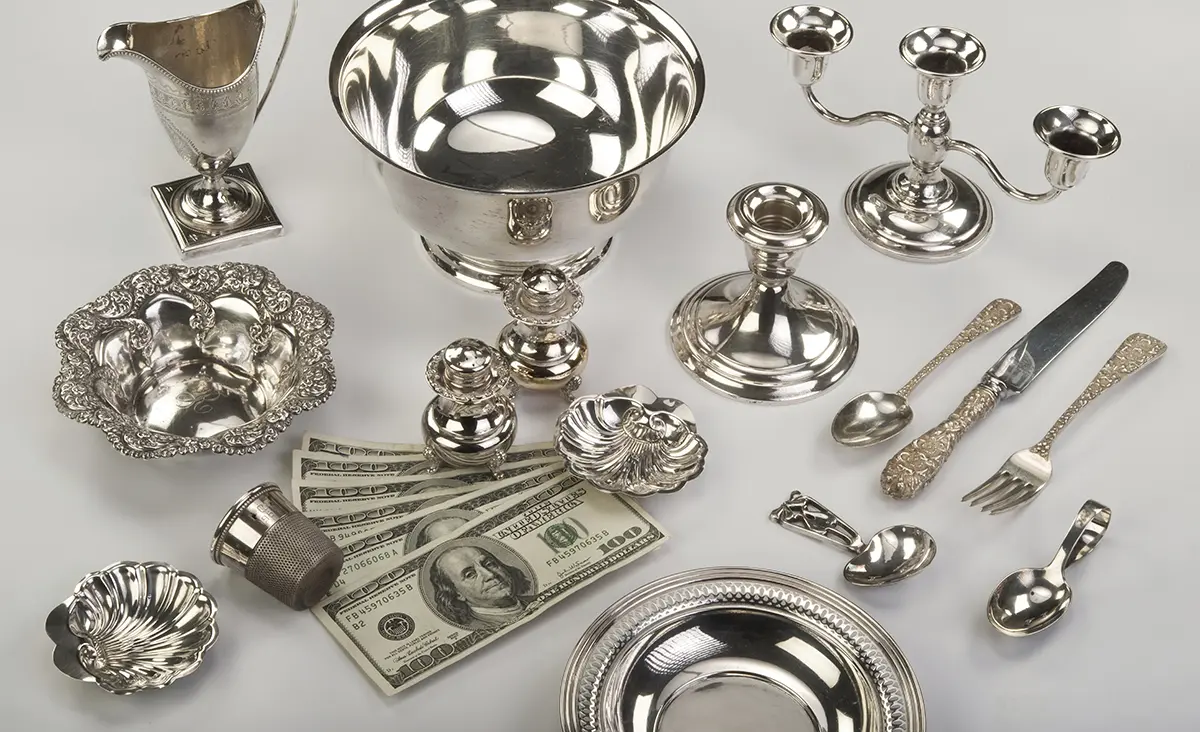 Sterling silver bowls, candlesticks, a creamer and ornate flatware laid out beside a fan of hundred-dollar bills.